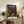 May include: Framed Spirited Away movie poster with Japanese text and Studio Ghibli artwork showing young girl in pink dress on traditional Japanese street, displayed on rustic wooden dresser beside brown leather chair, small potted fern, ceramic vases and books in warm-toned living room setting