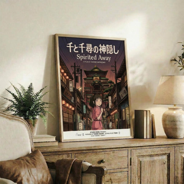 May include: Framed Spirited Away movie poster with Japanese text and Studio Ghibli artwork showing young girl in pink dress on traditional Japanese street, displayed on rustic wooden dresser beside brown leather chair, small potted fern, ceramic vases and books in warm-toned living room setting