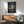 May include: Framed Spirited Away movie poster hanging on white wall above wooden sideboard with lamp and green upholstered chair. Poster features anime girl with dark hair in red clothing against twilight scene with train and spirits. Warm natural lighting creates shadows across minimalist room setting.