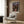 May include: Framed Spirited Away movie poster mounted on wood-paneled wall above modern cream armchair with geometric pillow. Poster shows young girl in pink outfit on traditional Japanese street with lanterns and buildings in twilight colors. Room features brass floor lamp, side table with open book, and neutral decor.