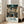 May include: Framed movie poster leaning against white wall next to beige sofa in bright living room with windows. Poster shows anime-style girl in traditional Japanese clothing carrying wooden bucket through bustling marketplace with lanterns and food stalls. Rich teal and orange colors throughout detailed street scene. Japanese and English text at bottom reads "Spirited Away" with Studio Ghibli credits.