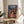 May include: Framed Spirited Away movie poster featuring animated girl in red outfit standing on wooden bridge with ornate Japanese bathhouse building behind her. Poster displays Japanese text and "Spirited Away" title against twilight sky. Frame sits on round wooden side table next to beige upholstered chair and plant leaves in bright, minimalist room setting.