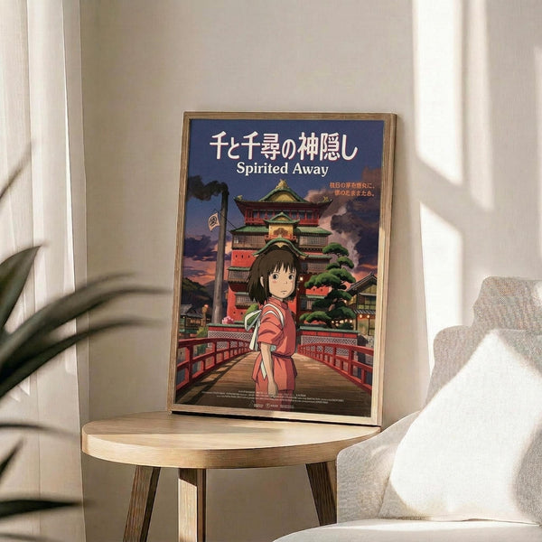 May include: Framed Spirited Away movie poster featuring animated girl in red outfit standing on wooden bridge with ornate Japanese bathhouse building behind her. Poster displays Japanese text and "Spirited Away" title against twilight sky. Frame sits on round wooden side table next to beige upholstered chair and plant leaves in bright, minimalist room setting.