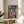 May include: Framed Spirited Away movie poster featuring Chihiro in pink dress against mystical Japanese bathhouse backdrop, displayed on wooden round side table next to beige upholstered chair with decorative plant nearby. Natural sunlight casts shadows across neutral-toned room setting. Poster shows detailed anime artwork with Japanese text and "Studio Ghibli Presents" branding in traditional illustration style.