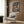 May include: Framed Spirited Away movie poster mounted on wood-paneled wall above beige upholstered armchair with geometric patterned pillow. Poster features anime character with reddish-brown hair in traditional clothing against fantastical Japanese architecture background with Japanese and English text. Modern reading nook setting with golden floor lamp and dark side table with open book.