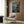 May include: Framed Studio Ghibli Spirited Away movie poster displayed on wood-paneled wall above beige armchair with geometric throw pillow. Poster shows young girl on wooden boat dock with ornate bathhouse building across teal waters. Modern living room setting with gold floor lamp and side table with open book. Warm neutral tones throughout scene.