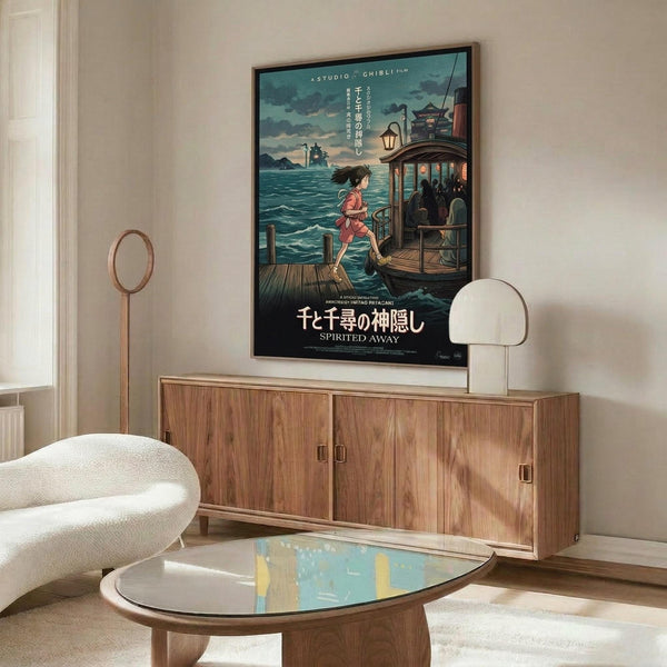 May include: Framed Spirited Away movie poster displayed above a wooden credenza in a modern living room. The poster shows an anime girl in pink running on wooden planks beside a traditional bathhouse over water, with Japanese text and "Studio Ghibli Film" at top. White sculptural chair and glass coffee table visible in foreground, creating a minimalist Scandinavian-style interior scene.