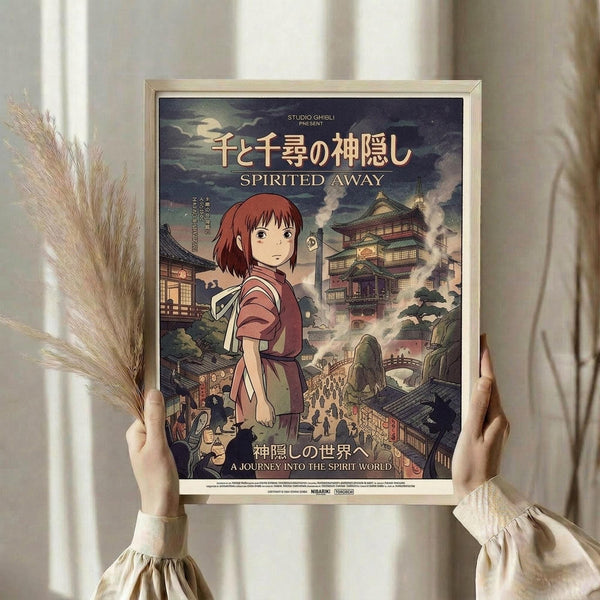 May include: Person in cream clothing holding framed Spirited Away movie poster featuring red-haired girl character against backdrop of Japanese buildings and bathhouse. Golden frame, dried pampas grass visible on left side. Soft natural lighting creates shadows on neutral wall background.