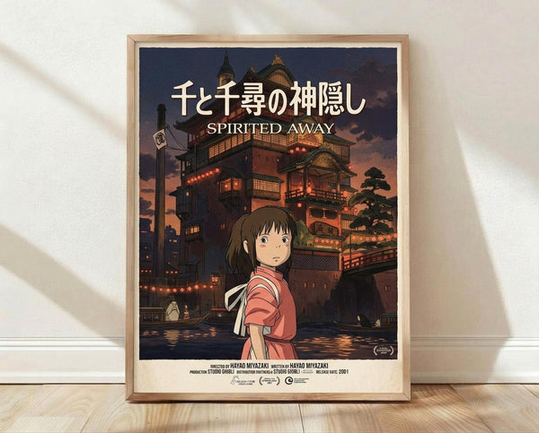 May include: Framed Spirited Away movie poster featuring young girl in red outfit standing before illuminated Japanese bathhouse at dusk. Traditional wooden architecture with lanterns and warm orange glow reflected in water. Poster displayed in light wood frame leaning against white wall on wooden floor with soft natural lighting and shadows.