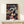 May include: Framed Spirited Away movie poster featuring main character Chihiro in pink kimono surrounded by animated characters including No-Face, Haku the dragon, and Zeniba, displayed on light wood shelf with small wrapped gift, against cream wall with natural lighting