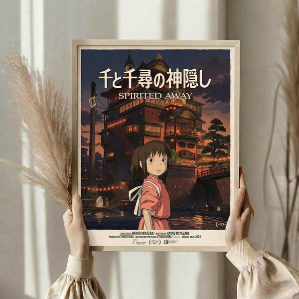 May include: Hands holding a framed Spirited Away movie poster featuring the young girl Chihiro in a pink shirt standing before an illuminated traditional Japanese bathhouse at twilight. The poster has Japanese text at the top with "Spirited Away" below, set in a light wood frame. Dried wheat stalks visible on the left side, neutral curtained background.