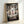 Framed poster of Studio Ghibli's Spirited Away featuring the main character Chihiro standing beneath a traditional Japanese torii gate. The artwork shows various spirit characters including No-Face, dragons, and magical creatures surrounding the young girl in a mystical forest setting with glowing lanterns. The poster displays both Japanese text and "SPIRITED AWAY" in English at the bottom, all contained within a light wooden frame leaning against a white wall.