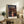 May include: Framed Spirited Away movie poster featuring young girl in red striped shirt against twilight bathhouse backdrop, displayed on rustic wooden sideboard alongside fern plant, books, and ceramic vase in cozy living room with beige armchair and neutral decor