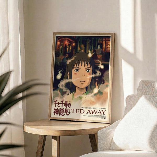 May include: Framed Spirited Away movie poster featuring animated character Chihiro surrounded by white spirits, with colorful bathhouse scene in background. Japanese and English text visible at bottom. Wooden frame sits on light wood round side table next to gray upholstered chair and plant leaves, with natural window lighting creating soft shadows on white wall.