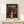 May include: Framed Spirited Away movie poster displayed on light wood shelf against white wall. Poster shows anime girl in pink shirt standing before illuminated traditional Japanese bathhouse at twilight. Small white gift box with twine sits in front of frame. Warm lighting creates cozy atmosphere.