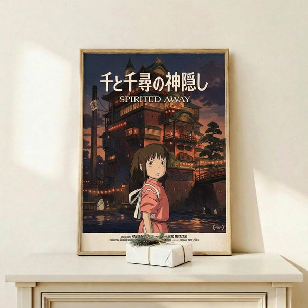 May include: Framed Spirited Away movie poster displayed on light wood shelf against white wall. Poster shows anime girl in pink shirt standing before illuminated traditional Japanese bathhouse at twilight. Small white gift box with twine sits in front of frame. Warm lighting creates cozy atmosphere.