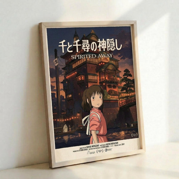 May include: Framed Spirited Away movie poster in light wood frame leaning against white wall. Features animated girl in pink top with Japanese bathhouse building behind her at twilight. Poster shows Japanese text at top with "SPIRITED AWAY" below in white letters. Rich sunset colors of orange, purple and deep blue create atmospheric background. Small production credits visible at bottom of poster.