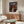 May include: Framed Spirited Away movie poster mounted on wall above wooden credenza in modern living room. Poster shows young girl in pink shirt against backdrop of ornate Japanese bathhouse at twilight. Room features white upholstered chair, glass-top oval coffee table, and warm wood furnishings. Natural lighting creates soft shadows across neutral-toned space.