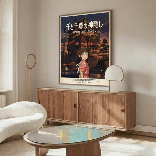 May include: Framed Spirited Away movie poster mounted on wall above wooden credenza in modern living room. Poster shows young girl in pink shirt against backdrop of ornate Japanese bathhouse at twilight. Room features white upholstered chair, glass-top oval coffee table, and warm wood furnishings. Natural lighting creates soft shadows across neutral-toned space.