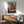 May include: Framed anime poster showing young girl in pink kimono leaping in front of ornate multi-story Japanese bathhouse with traditional curved roofs and red bridge. Poster features Japanese text at bottom. Displayed above mid-century modern wooden sideboard with white ceramic lamp in minimalist room setting with sage green upholstered chair and natural lighting casting geometric shadows on white wall.