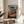 May include: Framed anime poster displayed on round wooden side table next to beige upholstered chair and potted plant. The colorful artwork shows a young girl in pink robes leaping through the air above a traditional multi-story Japanese bathhouse with green roofs, set against a blue sky with puffy clouds. Japanese text appears at the bottom of the poster in a thin natural wood frame.