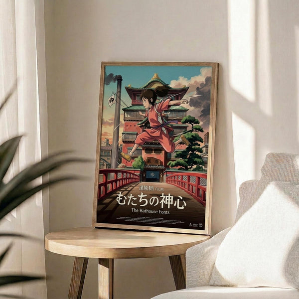 May include: Framed anime poster displayed on round wooden side table next to beige upholstered chair and potted plant. The colorful artwork shows a young girl in pink robes leaping through the air above a traditional multi-story Japanese bathhouse with green roofs, set against a blue sky with puffy clouds. Japanese text appears at the bottom of the poster in a thin natural wood frame.