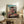 May include: Framed anime poster showing young girl in pink kimono leaping across traditional Japanese bathhouse with multi-tiered pagoda architecture, red bridge, and smokestack against cloudy sky. Japanese text at bottom. Displayed on rustic wooden console with books, ceramic vase, and fern plant in cozy living room setting with beige armchair.