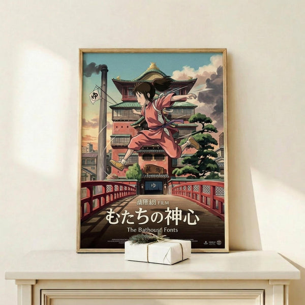 May include: Framed anime-style poster showing a young girl in pink kimono leaping over a red bridge with traditional Japanese bathhouse building in background. Artwork displays Studio Ghibli animation style with detailed architecture, cloudy sky, and Japanese text at bottom. Natural wood frame sits on light wooden shelf next to small white wrapped gift box against cream wall.