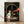May include: A framed movie poster featuring a young girl with brown hair in a white and green striped shirt standing in front of a stone archway tunnel. Red paper lanterns hang along a mysterious pathway leading into darkness. The poster displays Japanese text at the top and "Spirited Away" at the bottom. The wooden frame sits on light wooden flooring against a white wall with soft natural lighting casting gentle shadows.