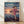 May include: Framed movie poster leaning against white wall on wooden floor. Features animated girl with brown hair in pink outfit running on flooded train tracks toward sunset. Train visible in background across water. Japanese and English text at top reads "Spirited Away." Directed by Hayao Miyazaki and Studio Ghibli credits at bottom. Natural wood frame, soft lighting creates shadows on wall.