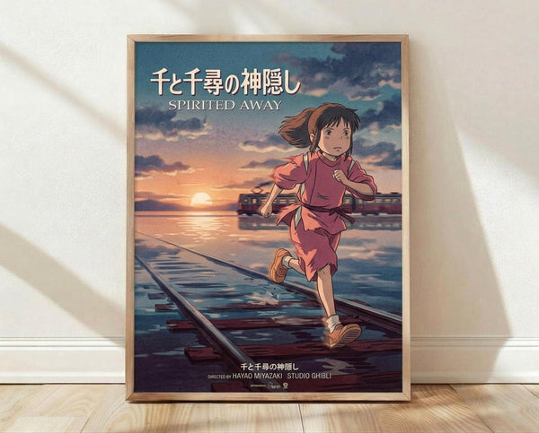 May include: Framed movie poster leaning against white wall on wooden floor. Features animated girl with brown hair in pink outfit running on flooded train tracks toward sunset. Train visible in background across water. Japanese and English text at top reads "Spirited Away." Directed by Hayao Miyazaki and Studio Ghibli credits at bottom. Natural wood frame, soft lighting creates shadows on wall.