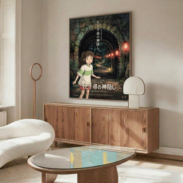 May include: Framed Spirited Away movie poster mounted on white wall above wooden credenza in modern living room. Poster features young girl character Chihiro standing before stone archway tunnel with red lanterns. Room includes white curved chair, oval glass coffee table, and floor lamp. Warm natural lighting creates shadows on furniture.