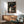 May include: Framed Spirited Away movie poster hanging on white wall above wooden sideboard with table lamp. Green upholstered armchair with wooden arms sits nearby. Poster shows young girl character standing in stone archway tunnel with red lanterns, featuring Japanese text and "Spirited Away" title. Warm sunlight streams across minimalist room with mid-century modern furniture.