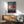 May include: Framed Spirited Away movie poster mounted on white wall above wooden sideboard with table lamp. Shows animated girl character running on wooden planks over water at sunset with Japanese text and "Spirited Away" title. Natural wood frame matches mid-century modern furniture including sage green upholstered armchair. Warm sunlight creates shadows across minimalist living room setting.