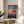May include: Framed Spirited Away movie poster featuring animated girl with brown hair in pink outfit running on wooden railway tracks over water at sunset. Poster displays Japanese text and "SPIRITED AWAY" title with Studio Ghibli credits. Light wooden frame sits on round wooden side table next to beige upholstered chair and green plant leaves in bright, minimalist room setting.