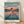 May include: Framed poster of anime character Chihiro from Spirited Away standing on flooded railway tracks. The young girl wears a pink outfit and looks back over her shoulder. Behind her stretches a dreamlike landscape with mountains, turquoise water, and sunset sky in soft pastels. Japanese and English text at top reads "Spirited Away." Light wooden frame sits on polished floor against white wall with soft shadows.