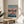 May include: Framed Spirited Away movie poster displaying Chihiro in pink dress walking on flooded train tracks at sunset, with Japanese and English text at top. Wooden frame sits on round wooden side table next to beige upholstered chair and plant, with natural sunlight streaming through white curtains creating soft shadows on cream walls.