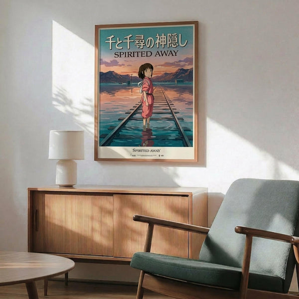 May include: Framed Spirited Away movie poster hanging on white wall above wooden sideboard with table lamp. Poster shows young girl in pink dress standing on flooded railway tracks with mountains in background, featuring Japanese and English text. Mid-century modern furniture including teal upholstered armchair with wooden arms visible in foreground. Natural sunlight creates geometric shadows across the minimalist interior scene.