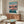 May include: Spirited Away movie poster mockup in modern living room setting with wooden sideboard, white upholstered chair, glass coffee table, and white table lamp. Poster shows anime character Chihiro in pink dress standing on flooded railway tracks with mountains and sunset sky in background. Japanese and English text at top. Warm wood tones throughout room with cream walls and natural lighting.