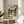 May include: Framed Spirited Away movie poster featuring anime girl with brown hair in green and white striped shirt standing in traditional Japanese garden setting with stone lanterns and pond. Poster displayed on round wooden side table next to beige upholstered chair with white throw pillow. Natural sunlight creates shadows on neutral wall behind frame.