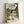 May include: Framed poster print of animated film Spirited Away featuring young girl with brown bob haircut in green and white striped shirt standing in traditional Japanese courtyard with stone lanterns, small stream, lush green trees, and traditional buildings with tiled roofs. Light wood frame leaning against white wall. Japanese and English text at top reads "Spirited Away - A Journey Beyond the Ordinary."