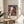 May include: Framed Spirited Away movie poster displayed on a round wooden side table next to a beige upholstered chair. The poster shows the main character Chihiro holding a paper lantern against a backdrop of the elaborate multi-story bathhouse building with traditional Japanese architecture. Soft natural light filters through white curtains, casting gentle shadows across the cream-colored wall. A green plant is partially visible in the corner, creating a cozy living room atmosphere.
