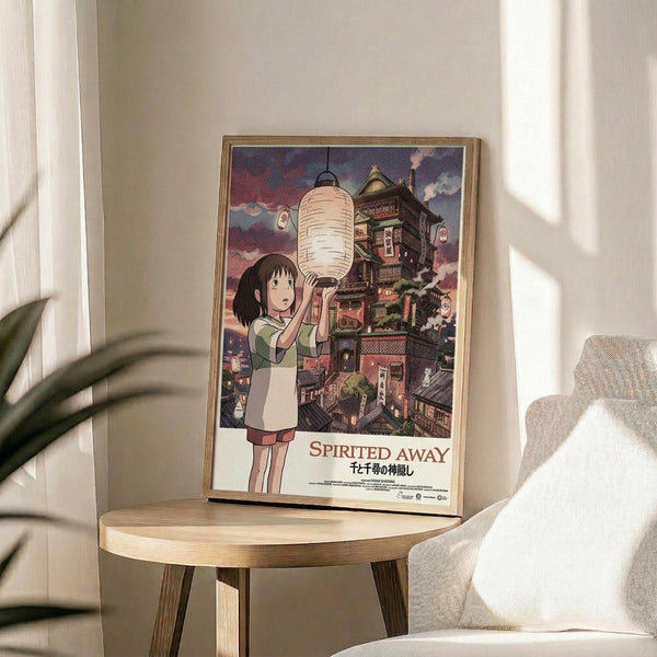 May include: Framed Spirited Away movie poster displayed on a round wooden side table next to a beige upholstered chair. The poster shows the main character Chihiro holding a paper lantern against a backdrop of the elaborate multi-story bathhouse building with traditional Japanese architecture. Soft natural light filters through white curtains, casting gentle shadows across the cream-colored wall. A green plant is partially visible in the corner, creating a cozy living room atmosphere.