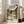 May include: Framed Spirited Away movie poster leaning against white wall next to beige armchair in bright living room. Poster shows anime girl with dark hair in green and white striped shirt standing in traditional Japanese garden setting with stone lanterns, pond, and wooden buildings. Light wood frame, natural lighting from windows.