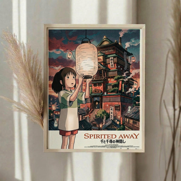May include: Framed Spirited Away movie poster displayed on white wall next to dried pampas grass. The artwork shows Chihiro holding a paper lantern with the elaborate bathhouse building in the background under a colorful sunset sky. The poster has Japanese text below the English title and is mounted in a light wooden frame.