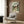 May include: Framed Spirited Away movie poster hanging on wood-paneled wall above beige armchair with geometric pillow. Poster shows young girl character Chihiro in green and white shirt against traditional Japanese buildings and garden setting. Modern reading nook with brass floor lamp and open book on side table.