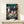 May include: Framed Spirited Away movie poster featuring young girl Chihiro surrounded by white spirits in traditional Japanese bathhouse setting with red lanterns and multi-story wooden architecture. Poster displayed on light wood shelf next to small white wrapped gift with twine and dried grass sprig against cream wall.