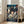 May include: Framed Studio Ghibli Spirited Away movie poster leaning against white wall in bright living room. Shows young girl with dark hair surrounded by ghostly spirits and traditional Japanese bathhouse architecture. Colorful anime artwork in light wood frame placed on hardwood floor next to beige couch with throw blanket. Natural sunlight streaming through windows creates warm, cozy home decor setting.