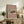 May include: Framed vintage Japanese woodblock print showing a traditional red temple gate building in snow, displayed on rustic wooden sideboard with fern plant, books, and ceramic vases. Poster has dusty rose background with architectural artwork featuring figures and cart near ornate temple structure. Natural lighting creates warm, cozy living room atmosphere.