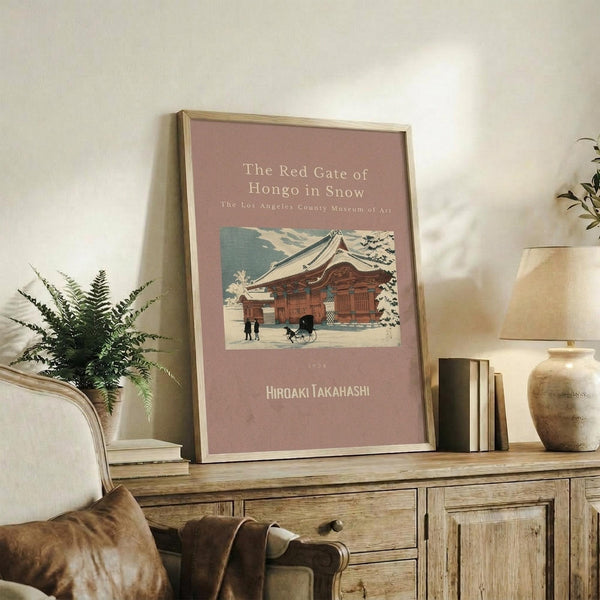 May include: Framed vintage Japanese woodblock print showing a traditional red temple gate building in snow, displayed on rustic wooden sideboard with fern plant, books, and ceramic vases. Poster has dusty rose background with architectural artwork featuring figures and cart near ornate temple structure. Natural lighting creates warm, cozy living room atmosphere.